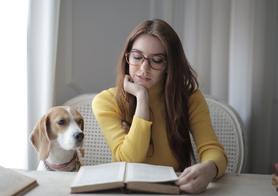 En kvinde med langt brunt hår og briller sidder ved et bord iført en gul striktrøje mens hun læser i en bog og kigger koncentreret ned på teksten en beagle står ved siden af hende og ser nysgerrigt på bogen