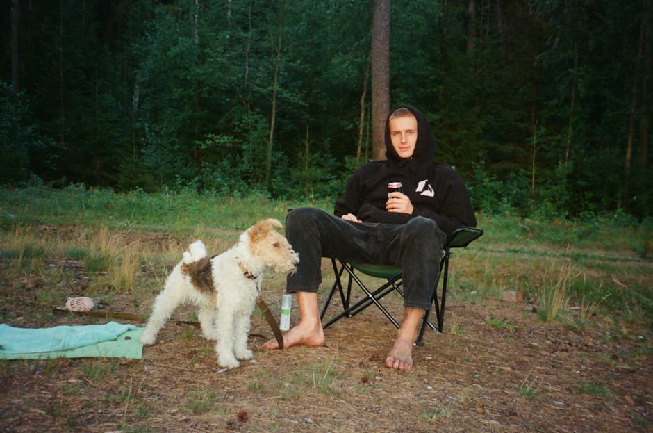 En mand sidder på en campingstol i skoven med barfødder og holder en dåse i hånden mens en fox-terrier står ved hans side på det græsklippede underlag