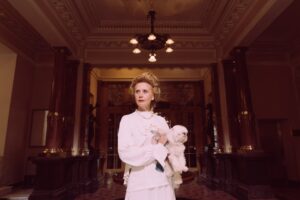 En kvinde i en elegant hvid kjole står i en rummelig foyer med højt loft og udsmykkede lys. Hun har en bølgende frisure og holder en lille hund tæt ind til sig. Bag hende er der marmorpiller og et kunstnerisk indretning, der tilføjer en vintage stemning til rummet.