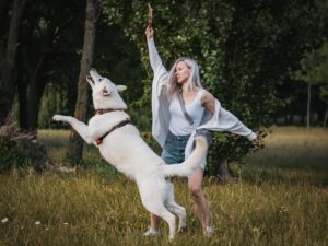 En kvinde med langt lyst hår leger med en hvid schweizisk hyrdehund i en grøn eng. Hunden står oprejst på bagbenene for at fange en gren, som kvinden holder op i luften. Begge ser glade ud, og der er træer i baggrunden.