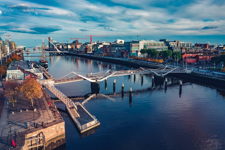 Billede af en bylandskab med en flod der løber gennem byen under en blå himmel med skyer broen med en moderne design strækker sig over vandet omgivet af bygninger i forskellige farver og træer med efterårsblade