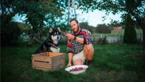 En mand knæler i en have og holder en skål med blommer, mens en norsk lundehund sidder ved siden af en trækasse i baggrunden omgiven af træer og en grønnere landskab.