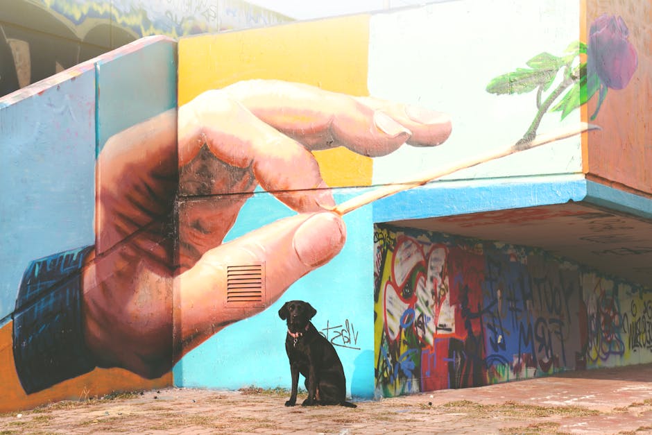 En sort hund sidder foran en farverig vægmaleri med en hånd, der holder en tandstik med en rose på. Væggen er prydet med graffiti i forskellige farver og mønstre.